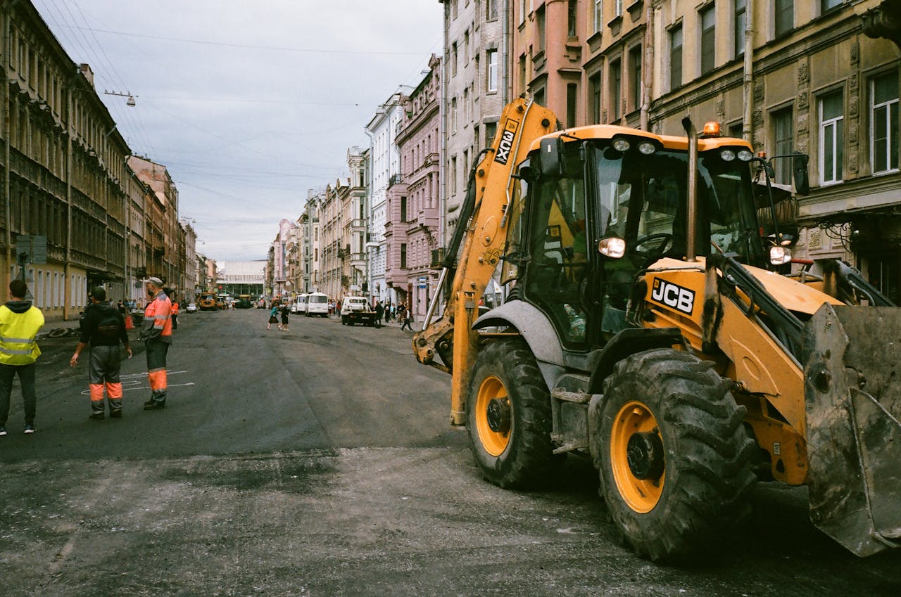 Teenused City street construction scene with workers and JCB excavator.